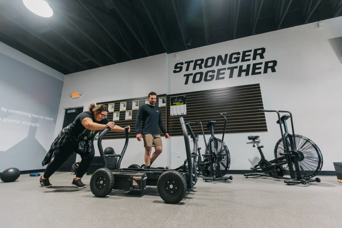 A member pushes a weighted sled across the training floor with a coach alongside at an Alloy Personal Training studio