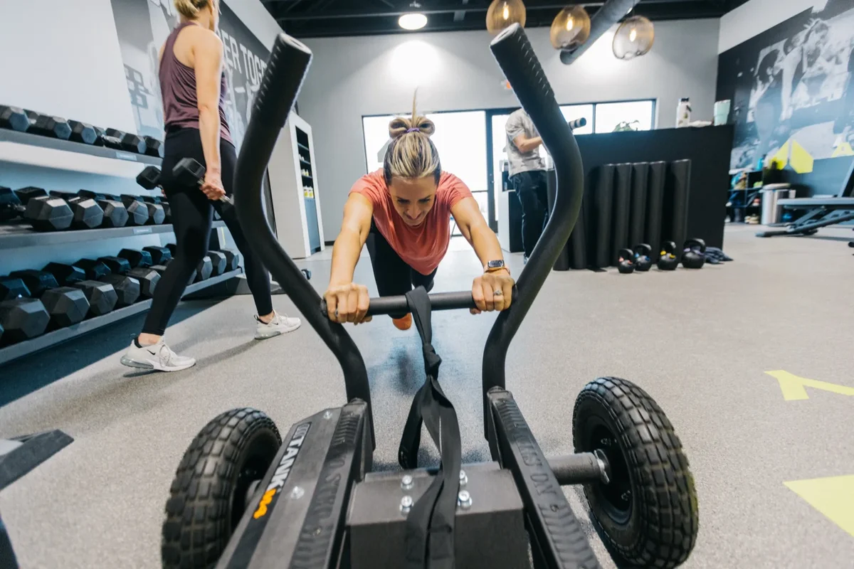 A member works through a sled push while a coach watches during a small group session at an Alloy Personal Training studio