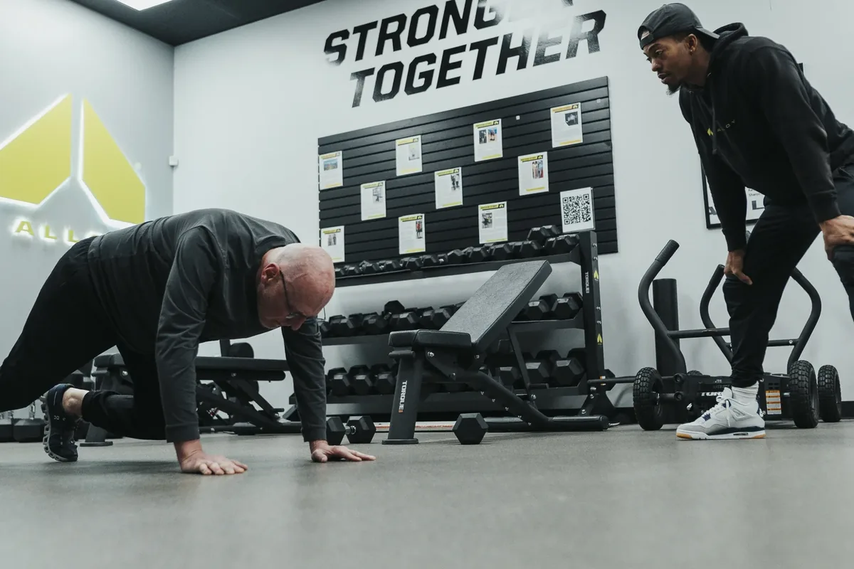 A coach watches an older member lean back into a suspension trainer hold during a session