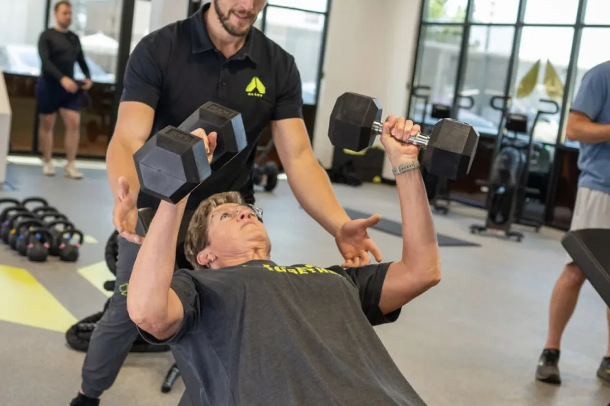 A coach spots an older member pressing dumbbells during a strength session at an Alloy Personal Training studio